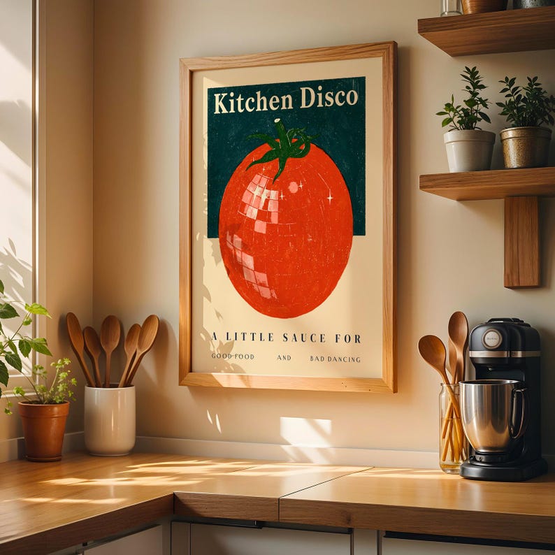 a framed poster of a tomato with a disco ball on it, hanging on a wall in a kitchen. The poster has the text "Kitchen Disco" and "A little sauce for good food and bad dancing".