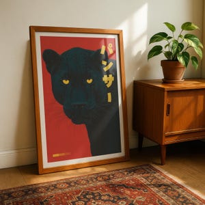 May include: A framed art print featuring a black panther on a red background. The panther's yellow eyes and Japanese text in gold add contrast. The print is in a wooden frame, leaning against a wall, with a rug and wooden cabinet in the background.