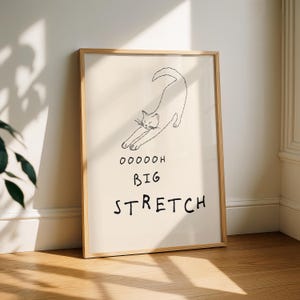 May include: A framed art print featuring a minimalist line drawing of a stretching cat. The artwork is in black and white, with the words "OOOOOH BIG STRETCH" below the cat illustration. The frame is a light wood color.