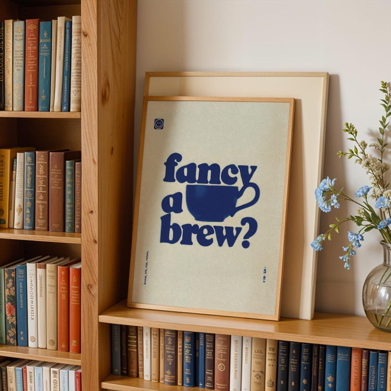 May include: A framed print with the words "fancy a brew?" in navy blue, with a navy blue coffee cup graphic. The print is in a light wood frame and is leaning against a larger, unframed print. The background is a light beige color.