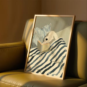 Puede incluir: Impresión artística enmarcada que representa a un perro blanco leyendo un libro bajo una manta a rayas. La obra de arte tiene un marco de madera clara y presenta rayas azules y blancas.