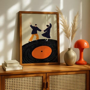 以下が含まれることがあります： レコードの上で踊る2人の人物を描いた、額入りのアートプリント。 このアートワークは、オレンジ色の中心を持つ黒いレコードを特徴とし、ダンサーはネイビーとオレンジ色で着飾っています。 プリントは木製のキャビネットに飾られています。