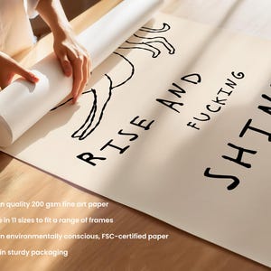 Op de afbeelding: Een opgerolde kunstprint op beige papier met de woorden "RISE AND FUCKING SHINE" in het zwart. Een zwarte lijntekening van een figuur staat boven de tekst. De print wordt door een persoon uitgerold.