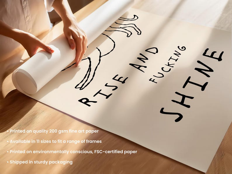 Op de afbeelding: Een opgerolde kunstprint op beige papier met de woorden "RISE AND FUCKING SHINE" in het zwart. Een zwarte lijntekening van een figuur staat boven de tekst. De print wordt door een persoon uitgerold.