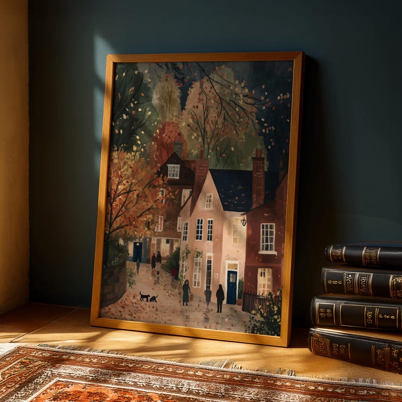 K&ouml;nnte beinhalten: Ein gerahmter Kunstdruck, der eine malerische Dorfszene mit Menschen zeigt, die auf einer Kopfsteinpflasterstra&szlig;e spazieren. Das Kunstwerk zeigt Geb&auml;ude mit wei&szlig;en und roten Backsteinfassaden, umgeben von B&auml;umen mit Herbstlaub. Der Rahmen ist hellbraun.