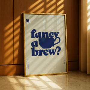 May include: A framed art print with a gold border. The print features the phrase "fancy a brew?" in dark blue, with a blue coffee cup graphic. The background is a light beige color. The print is displayed against a wooden wall.