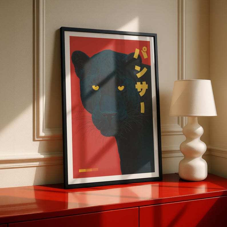 May include: Framed art print featuring a black panther illustration. The panther's silhouette is set against a red background, with yellow eyes and Japanese text. The artwork is in a black frame, displayed on a red cabinet.