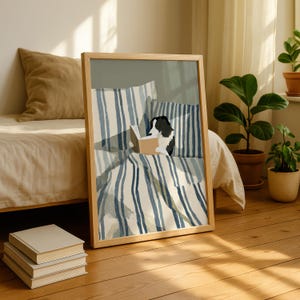 Op de afbeelding: Een ingelijste print toont een zwart-witte hond die een boek leest in bed. Het beddengoed heeft blauw-witte strepen. De print zit in een licht houten frame en leunt tegen een bed met een beige dekbed.