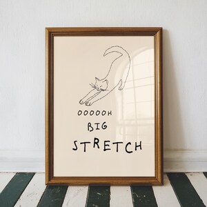 May include: A framed print featuring a minimalist line drawing of a stretching cat. The artwork is in black ink on a cream background, with the words "OOOOOH BIG STRETCH" below the cat. The frame is a warm brown color.