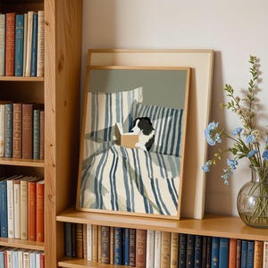 Op de afbeelding: Een ingelijste prent van een hond die in bed leest, met blauw-wit gestreept beddengoed. Het kunstwerk wordt tentoongesteld op een houten plank naast een boekenkast vol boeken en een vaas met blauwe bloemen.