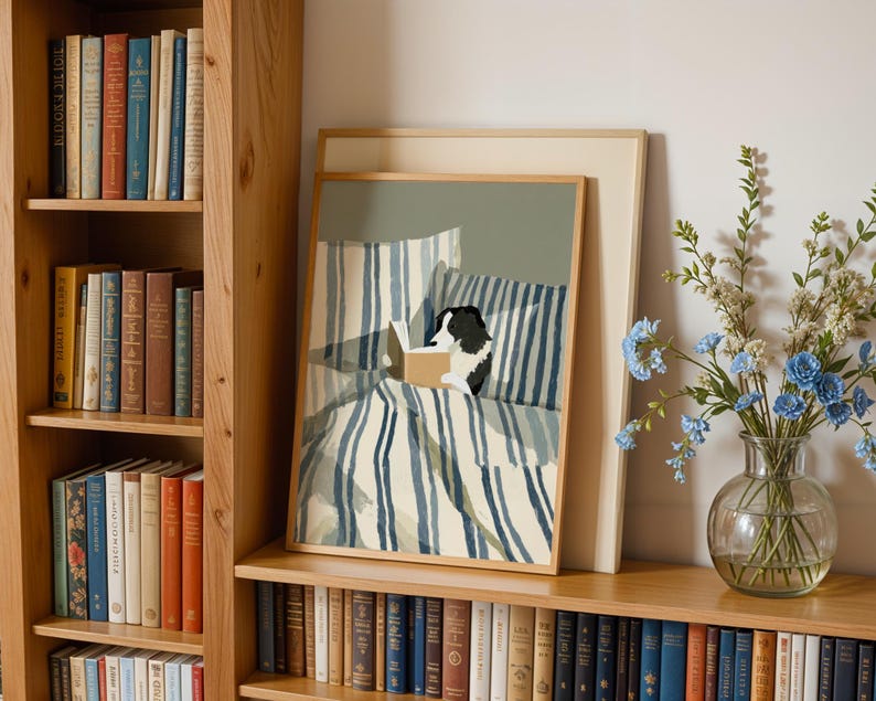 Op de afbeelding: Een ingelijste prent van een hond die in bed leest, met blauw-wit gestreept beddengoed. Het kunstwerk wordt tentoongesteld op een houten plank naast een boekenkast vol boeken en een vaas met blauwe bloemen.