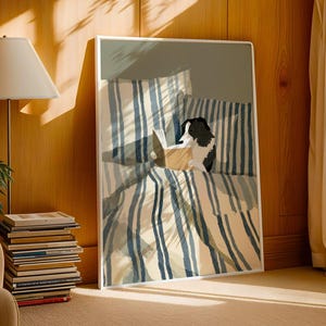 Op de afbeelding: Een kunstafdruk met een hond die in bed leest, met blauw-wit gestreepte beddengoed. Het kunstwerk staat in een witte lijst en leunt tegen een houten muur. Een stapel boeken en een lamp staan op de voorgrond.