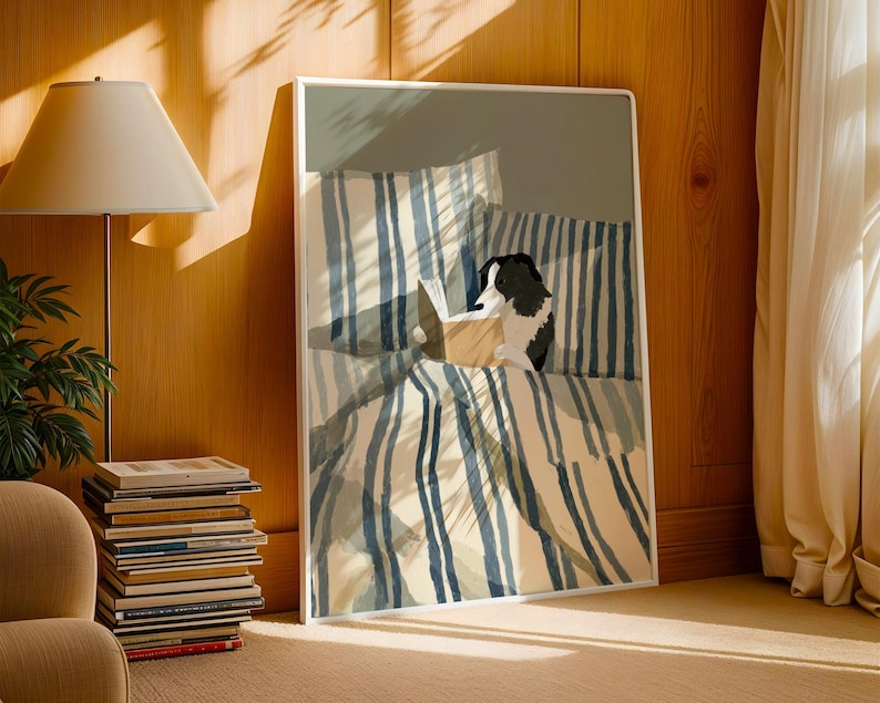 Op de afbeelding: Een kunstafdruk met een hond die in bed leest, met blauw-wit gestreepte beddengoed. Het kunstwerk staat in een witte lijst en leunt tegen een houten muur. Een stapel boeken en een lamp staan op de voorgrond.