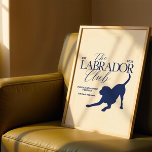Puede incluir: Un p&oacute;ster enmarcado con fondo beige claro y marco de madera. El p&oacute;ster presenta el texto "The Labrador Club" en azul marino, junto con una silueta de un perro Labrador. Tambi&eacute;n se incluye el texto "Puddle splashing forever" y "Eat hard, nap hard".