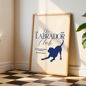 Puede incluir: Un p&oacute;ster enmarcado con fondo beige claro y texto y gr&aacute;fico azul marino. El texto dice "The Labrador Club" con la silueta de un perro labrador. El texto adicional incluye "Puddle splashing forever" y "Eat hard, nap hard."