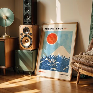 Framed Mount Fuji poster leaning on the floor beside vintage speakers, depicting iconic Japanese scenery with snowy peak, red sun, and crashing waves. Ideal Japanese decor or Mount Fuji print for modern interiors.