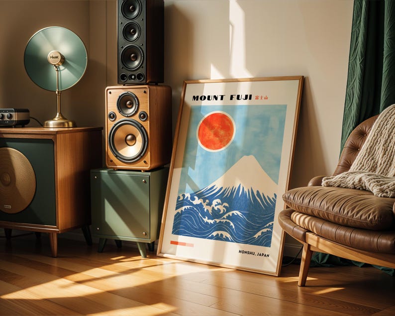 Framed Mount Fuji poster leaning on the floor beside vintage speakers, depicting iconic Japanese scenery with snowy peak, red sun, and crashing waves. Ideal Japanese decor or Mount Fuji print for modern interiors.