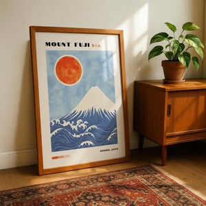 Framed Mount Fuji artwork leaning beside a wooden cabinet and potted plant on a patterned rug. Japanese wall art print with mountain, waves, and red sun—perfect for Japanese decor or mountain-themed gift.