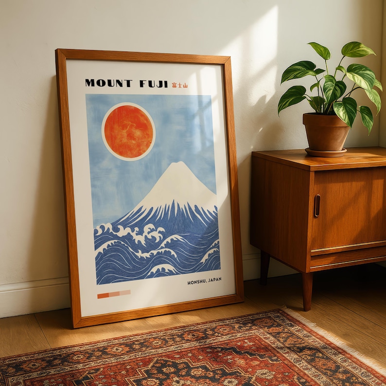 Framed Mount Fuji artwork leaning beside a wooden cabinet and potted plant on a patterned rug. Japanese wall art print with mountain, waves, and red sun—perfect for Japanese decor or mountain-themed gift.