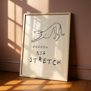 May include: A framed print with a black line drawing of a dog stretching. The text "OOOOOH BIG STRETCH" is below the dog illustration. The frame is a light beige colour, and the background is a cream colour.