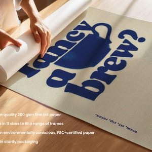 May include: A rolled-up art print featuring the text "Fancy a brew?" in blue, along with a blue coffee cup illustration. The print is on a beige background. A person's hands are visible, holding the rolled print.