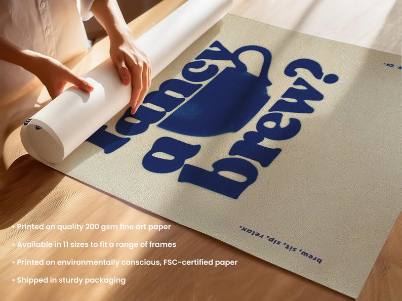 May include: A rolled-up art print featuring the text "Fancy a brew?" in blue, along with a blue coffee cup illustration. The print is on a beige background. A person's hands are visible, holding the rolled print.