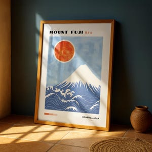 Mount Fuji print in wooden frame leaning against a deep blue wall on a tiled floor, next to a clay vase. Japanese poster showcases snowy mountain, red sun, and ocean waves in a bold, minimal style.