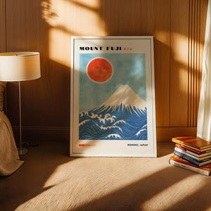 Mount Fuji print in a light wood frame resting on carpet beside a lamp and stack of books. Japanese artwork features mountain, ocean waves, and red sun in bold, minimalist style. Perfect Japanese wall art or gift.