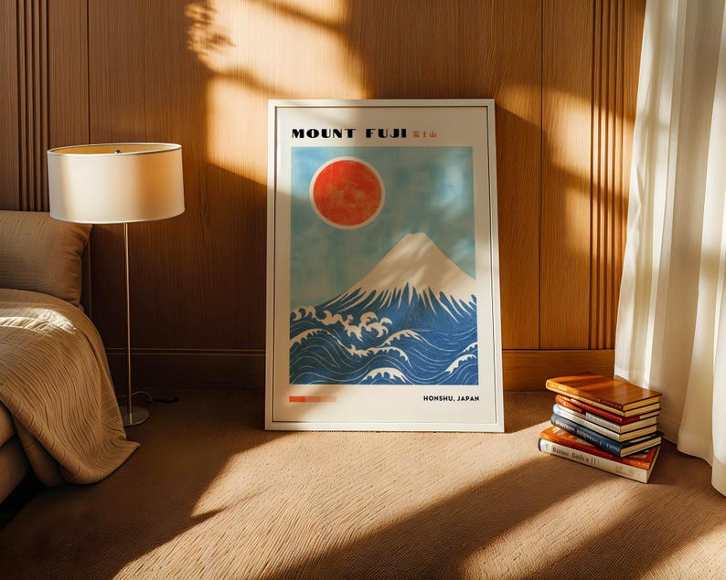 Mount Fuji print in a light wood frame resting on carpet beside a lamp and stack of books. Japanese artwork features mountain, ocean waves, and red sun in bold, minimalist style. Perfect Japanese wall art or gift.