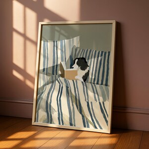 Op de afbeelding: Ingelijste kunstdruk met een zwart-witte hond die een boek leest in bed. Het beddengoed heeft blauw-witte verticale strepen. Het kunstwerk zit in een lichtgekleurde houten lijst en leunt tegen een muur met zonlicht.