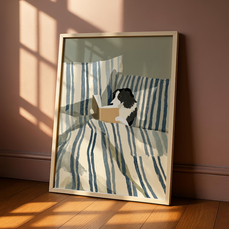 Op de afbeelding: Ingelijste kunstdruk met een zwart-witte hond die een boek leest in bed. Het beddengoed heeft blauw-witte verticale strepen. Het kunstwerk zit in een lichtgekleurde houten lijst en leunt tegen een muur met zonlicht.