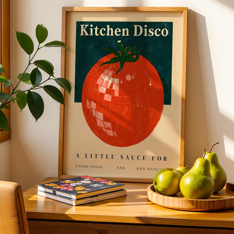 a framed poster of a tomato with a disco ball, placed on a wooden table next to a stack of books and a plate of green pears.
