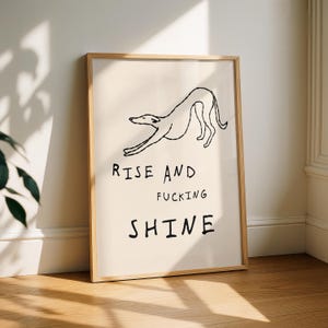 Op de afbeelding: Een ingelijste kunstdruk met een minimalistische lijntekening van een hond die zich uitrekt. De tekst "RISE AND FUCKING SHINE" is in vette, zwarte letters onder de hondenillustratie gedrukt. De lijst heeft een lichte houtkleur.