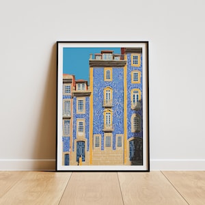 K&ouml;nnte beinhalten: Ein gerahmter Kunstdruck einer Geb&auml;udefassade mit blauen Fliesen und gelben Akzenten. Das Geb&auml;ude hat mehrere Fenster und T&uuml;ren, mit einem klaren blauen Himmel im Hintergrund. Der Druck wird auf einem Holzboden pr&auml;sentiert.