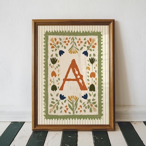 May include: Framed art print featuring the letter "A" in a rust-orange hue, surrounded by a floral border with green, blue, and yellow accents. The artwork is set against a cream background with vertical stripes, encased in a brown wooden frame.