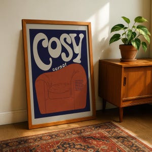 May include: A framed art print with a wooden frame. The print features the word "Cosy" in large white letters above an illustration of an orange armchair on a navy blue background. A potted plant sits on a wooden cabinet.