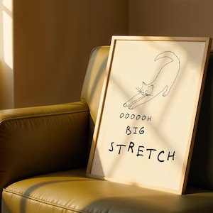 May include: A framed art print featuring a minimalist line drawing of a stretching cat. The artwork is on a cream-colored background with the words "Ooooooh Big Stretch" in black. The frame is a light wood color.