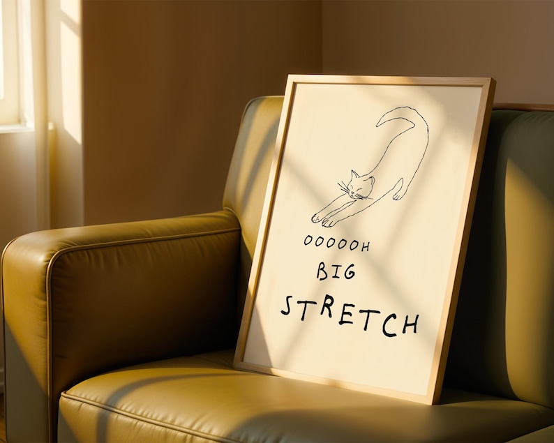 May include: A framed art print featuring a minimalist line drawing of a stretching cat. The artwork is on a cream-colored background with the words "Ooooooh Big Stretch" in black. The frame is a light wood color.