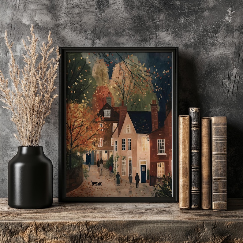 K&ouml;nnte beinhalten: Ein gerahmter Kunstdruck zeigt eine malerische Dorfansicht mit H&auml;usern in Beige- und Rott&ouml;nen, vor dem Hintergrund von Herbstlaub. Eine schwarze Vase mit getrockneten Pflanzen und ein Stapel alter B&uuml;cher stehen auf einem Holzregal.