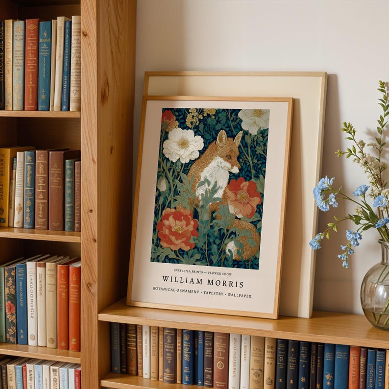 A framed print of a fox surrounded by flowers is displayed on a bookshelf, with a vase of blue flowers to the right.