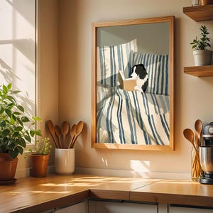 Op de afbeelding: Een ingelijst kunstwerk toont een zwart-witte hond die in bed leest. Het beddengoed heeft blauw-witte strepen. Het kunstwerk staat in een licht houten lijst, tentoongesteld op een aanrecht met planten en keukengerei.