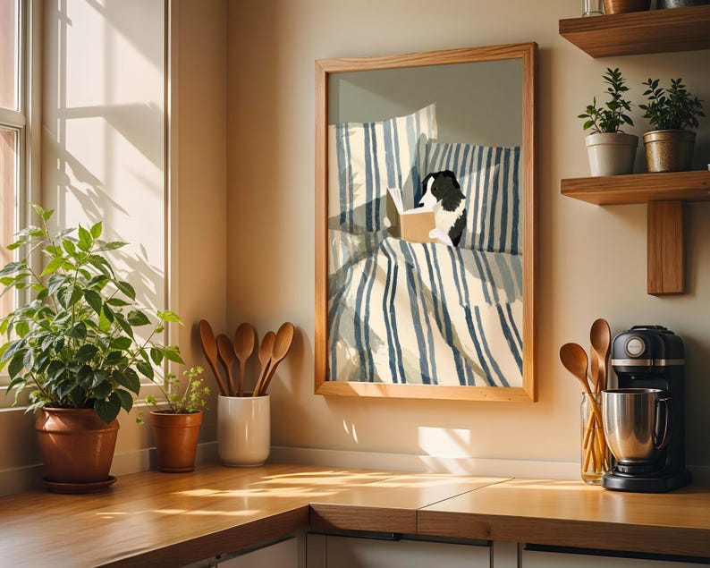 Op de afbeelding: Een ingelijst kunstwerk toont een zwart-witte hond die in bed leest. Het beddengoed heeft blauw-witte strepen. Het kunstwerk staat in een licht houten lijst, tentoongesteld op een aanrecht met planten en keukengerei.