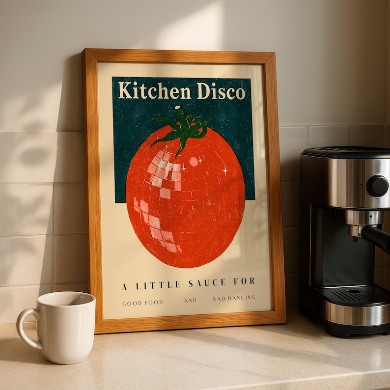 A framed poster with a tomato graphic and the text "Kitchen Disco" is displayed on a kitchen counter next to a coffee maker.