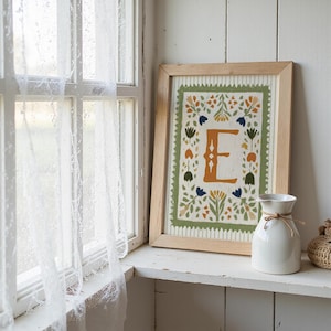 May include: A framed art print featuring the letter "E" in an orange hue, surrounded by floral and leaf designs in green, blue, and orange. The print is in a light wood frame. A white vase with a twine bow sits nearby on a white shelf.