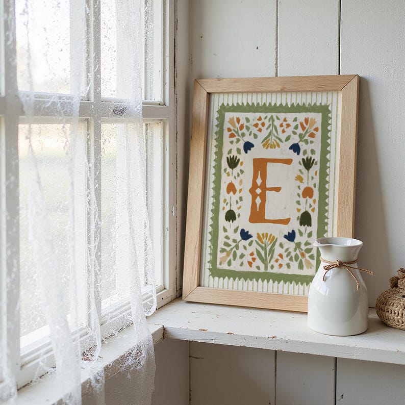May include: A framed art print featuring the letter "E" in an orange hue, surrounded by floral and leaf designs in green, blue, and orange. The print is in a light wood frame. A white vase with a twine bow sits nearby on a white shelf.