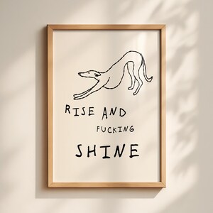 Op de afbeelding: Een ingelijste kunstprint met een zwarte lijntekening van een hond die zich uitrekt. De tekst "RISE AND FUCKING SHINE" staat in zwarte letters eronder. Het kunstwerk staat op een cr&egrave;me achtergrond en in een lichtbruine houten lijst.