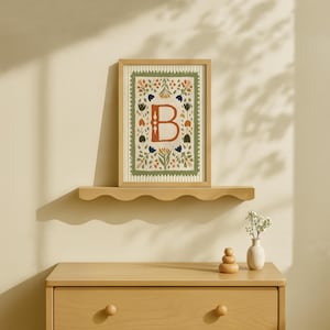 May include: A framed artwork featuring the letter "B" in a floral design. The artwork is in a wooden frame and rests on a scalloped shelf above a wooden dresser. The artwork has a cream background with a green border and colorful floral accents.