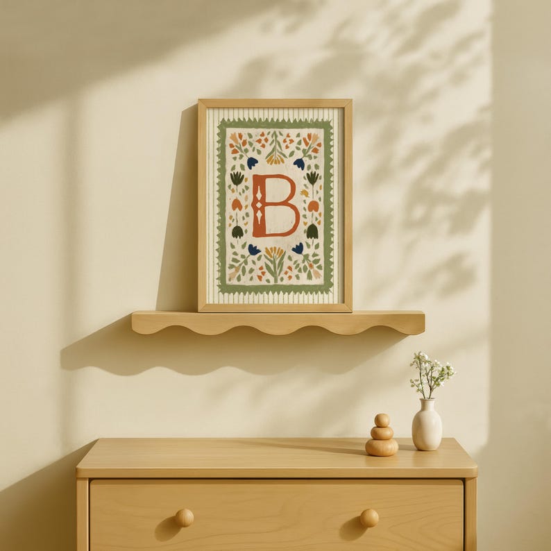May include: A framed artwork featuring the letter "B" in a floral design. The artwork is in a wooden frame and rests on a scalloped shelf above a wooden dresser. The artwork has a cream background with a green border and colorful floral accents.