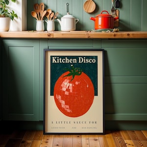 a framed poster with a large tomato graphic and text, placed on a kitchen counter with various kitchen utensils and appliances in the background.