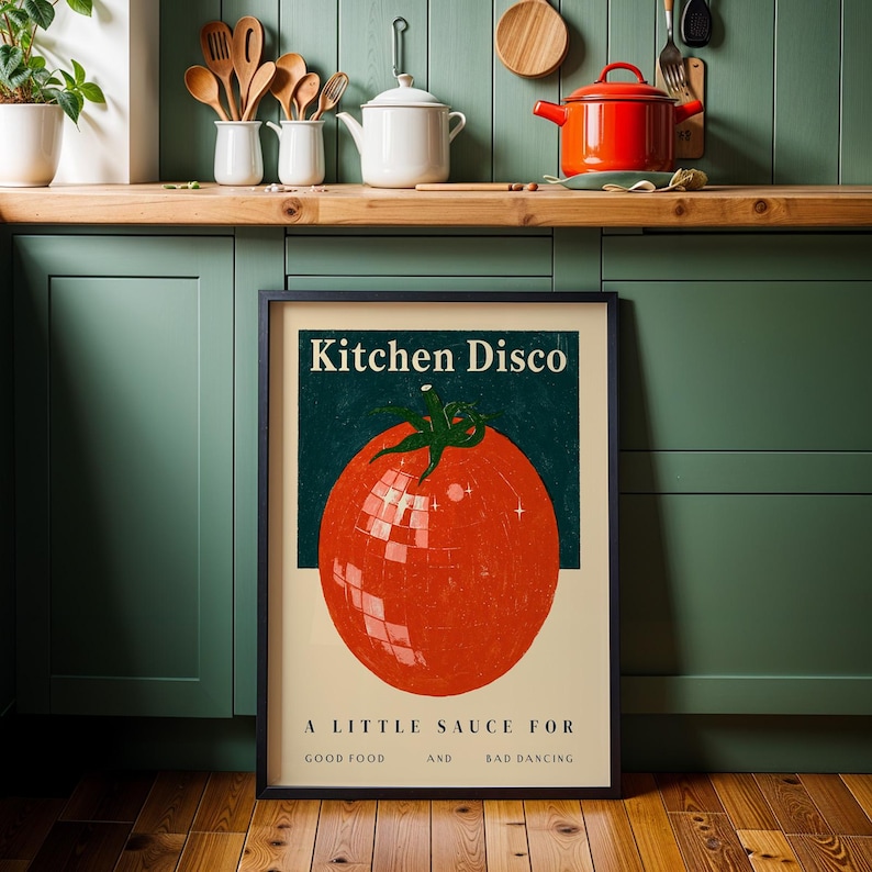 a framed poster with a large tomato graphic and text, placed on a kitchen counter with various kitchen utensils and appliances in the background.